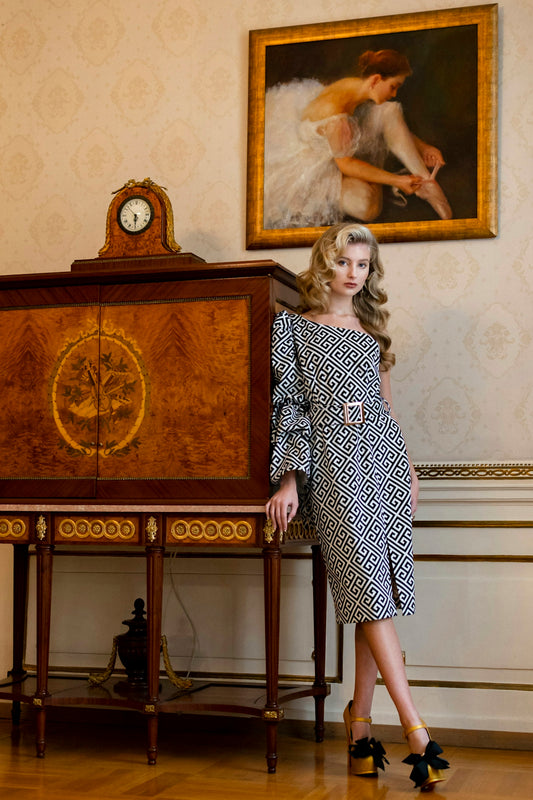 Elegant woman in patterned dress by antique cabinet and painting.