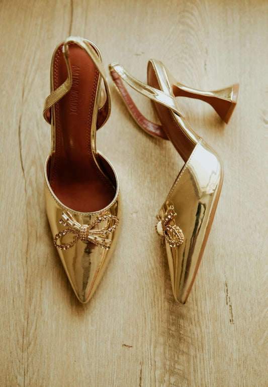 Two golden high-heeled shoes with rhinestone bows on wooden floor.